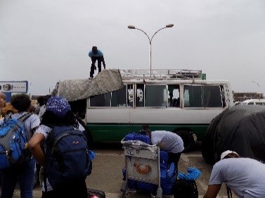 Drexel students, faculty and staff wearing backpacks stand in a parking lot in Senegal while a man covers a buss with a tarp. 